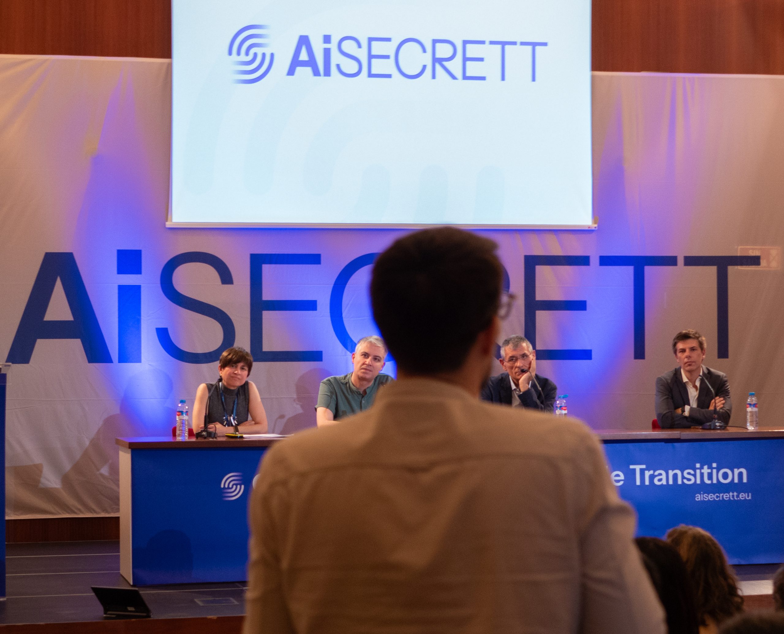 speaker addressing a panel during an AI-SECRETT event, with the project logo displayed on stage