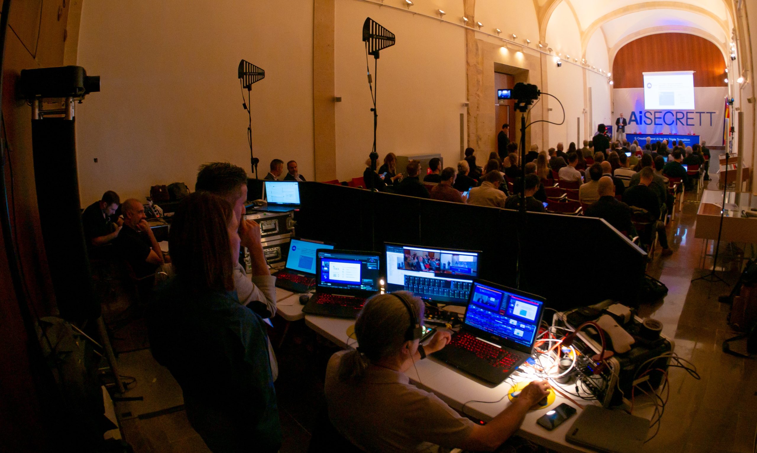 participants attend an AI-SECRETT event in an auditorium, with the production team operating audiovisual equipment in the foreground
