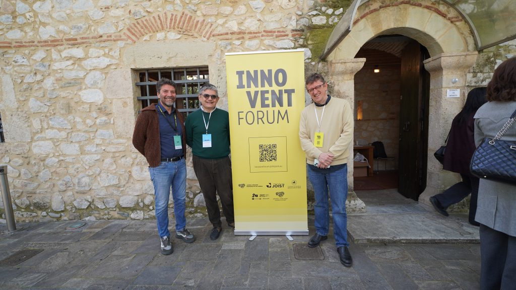 three participants posing outdoors next to an Innovent Forum banner outside the event venue in Larissa